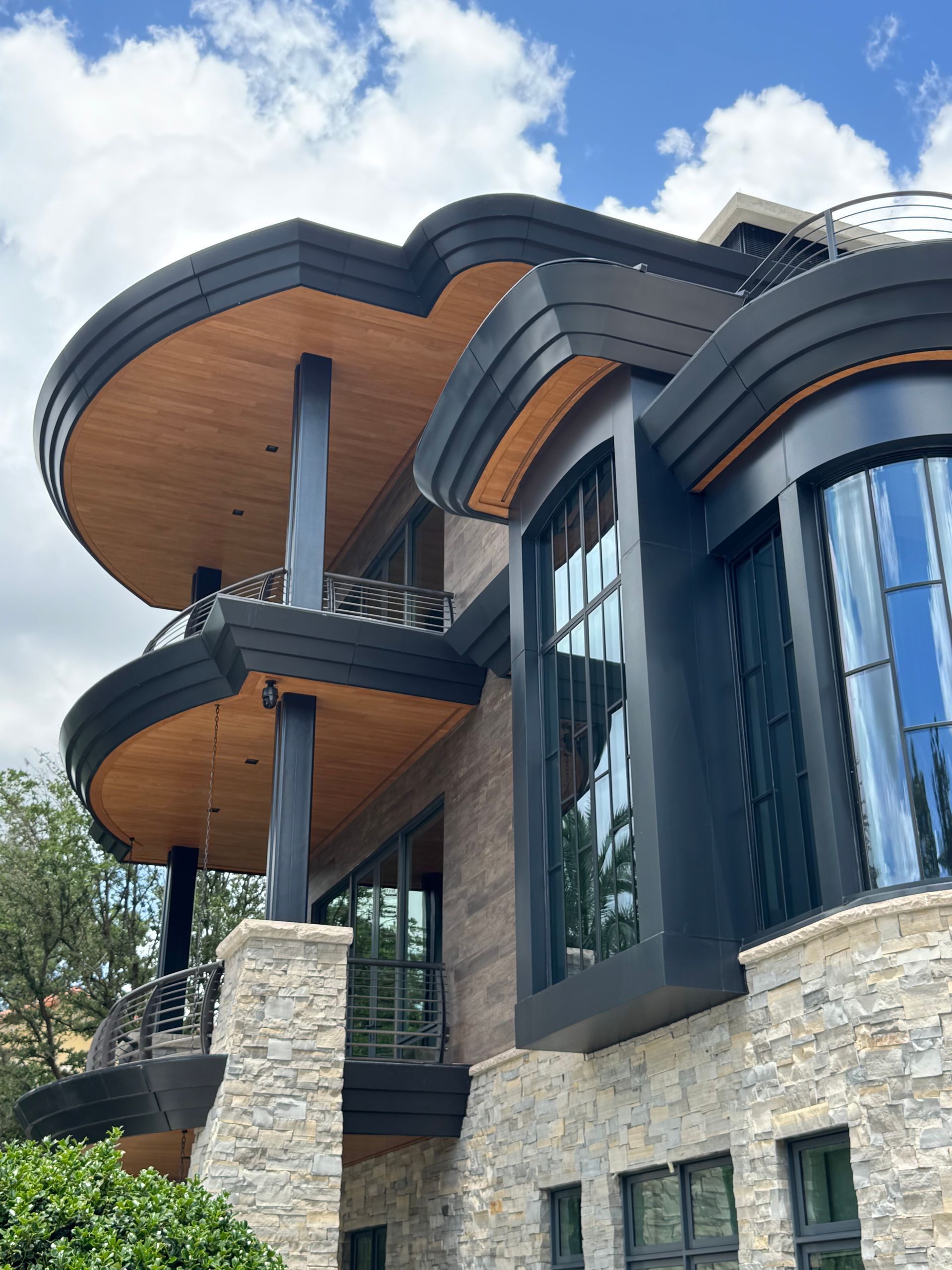 Modern house exterior with curved wooden balconies, dark trim, and stone walls under a blue sky