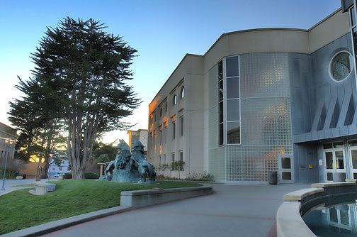 Modern building entrance with a curved canopy, glass facade, and landscaped walkway at sunset
