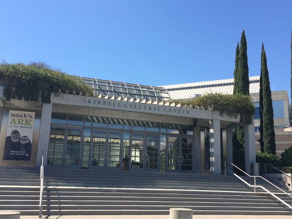 Airport curbside drop-off with white canopy, glass booth marked “C,” and a few people waiting