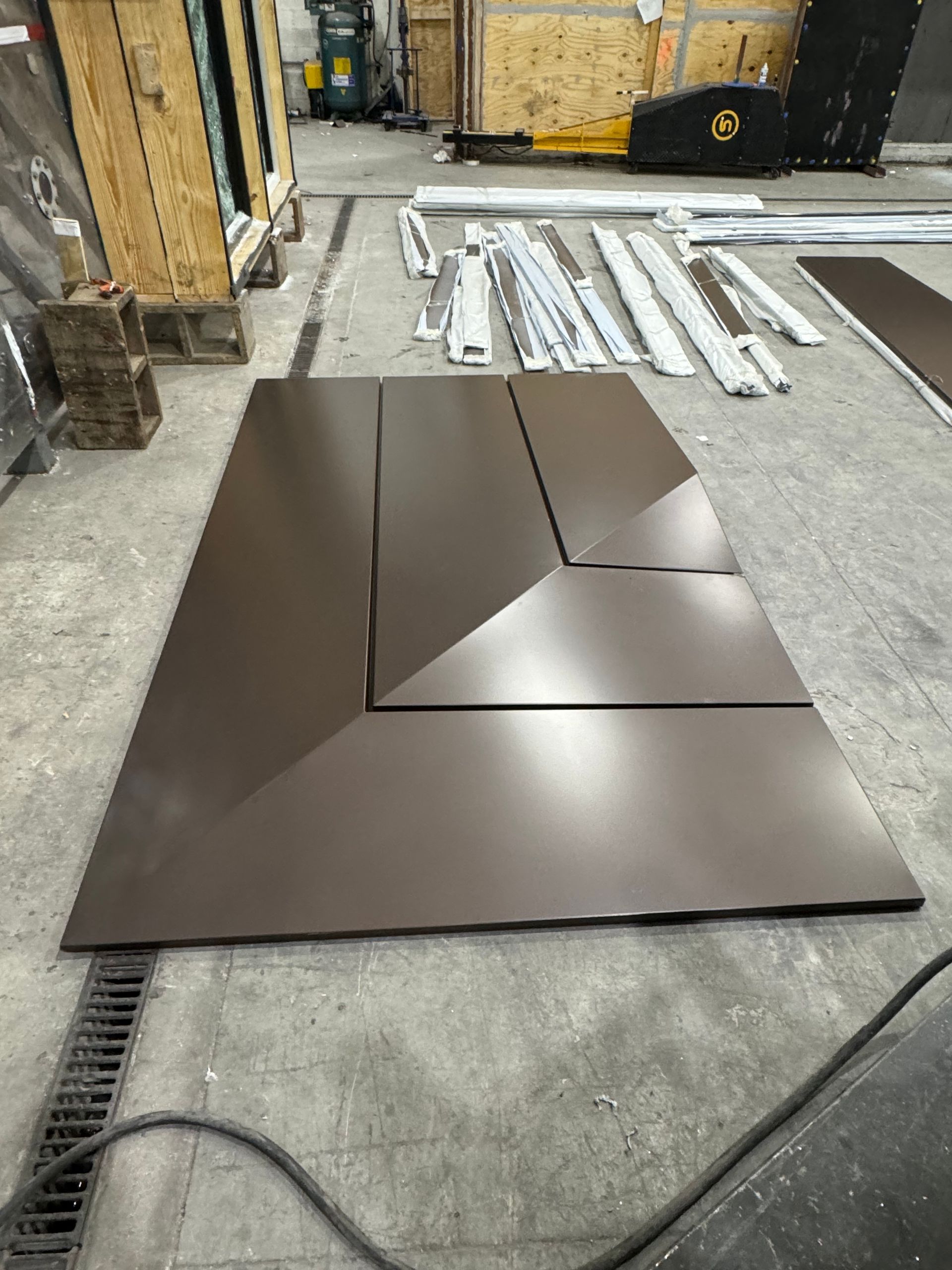 Dark brown geometric metal panel on a workshop floor with pipes and materials in the background