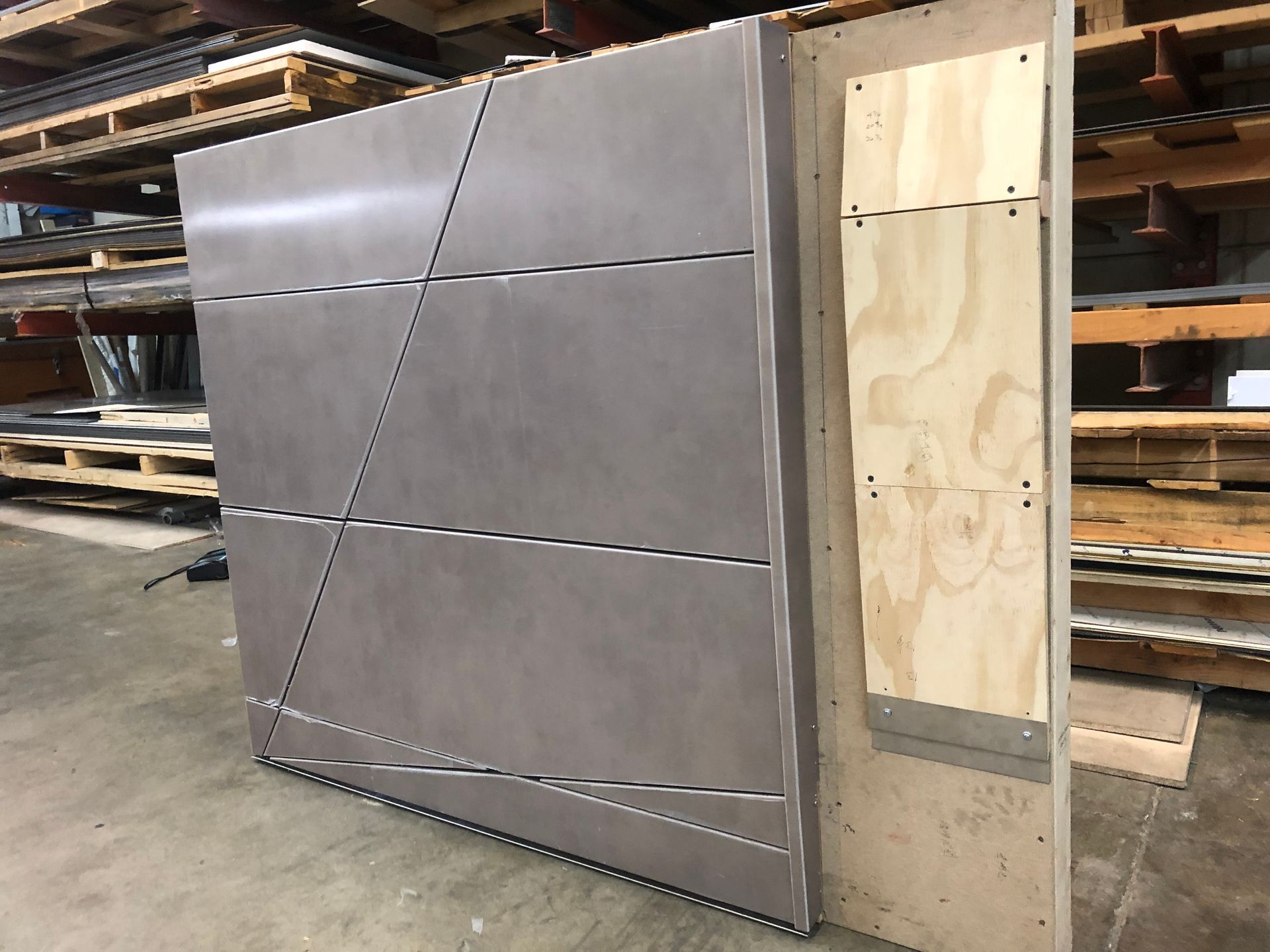 Large gray metal panel with diagonal seams leaning in a workshop beside a plywood frame