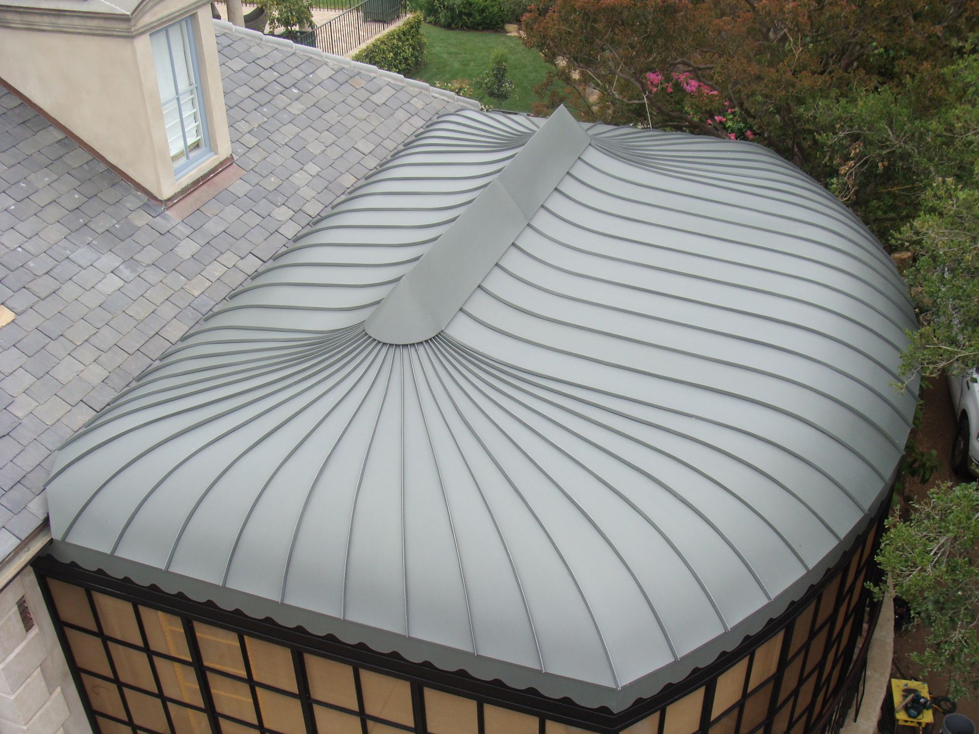 Curved gray metal roof with a central raised seam on a small rooftop structure beside a shingled house