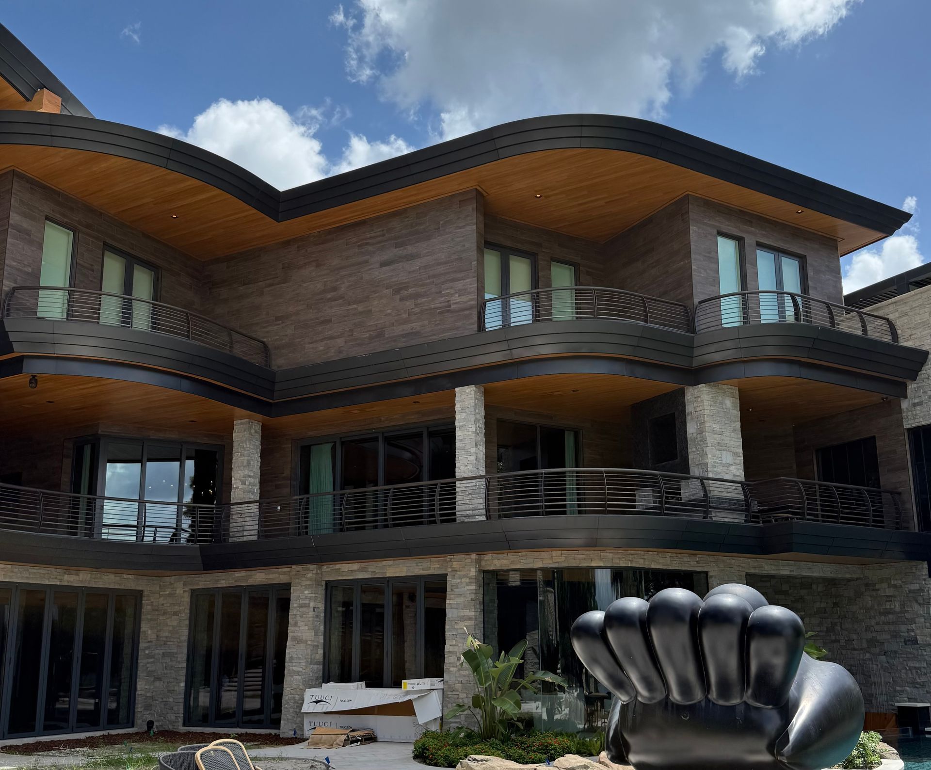 Modern multi-story house with curved wooden roofs and a large black sculpture in front under a blue sky