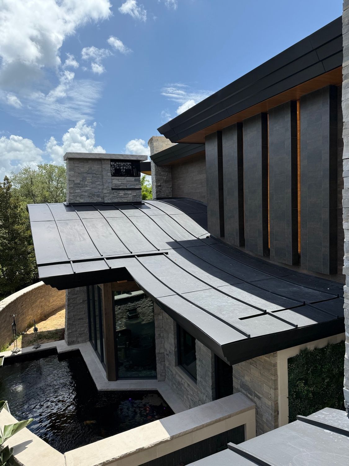 Modern house exterior with dark slate roof, stone walls, tall windows, and a small courtyard pool under a blue sky