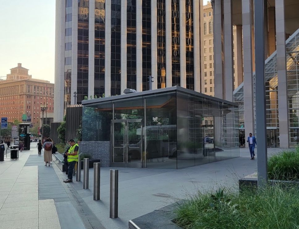 Glass transit kiosk on a city sidewalk beside tall office buildings, with pedestrians and a security guard nearby