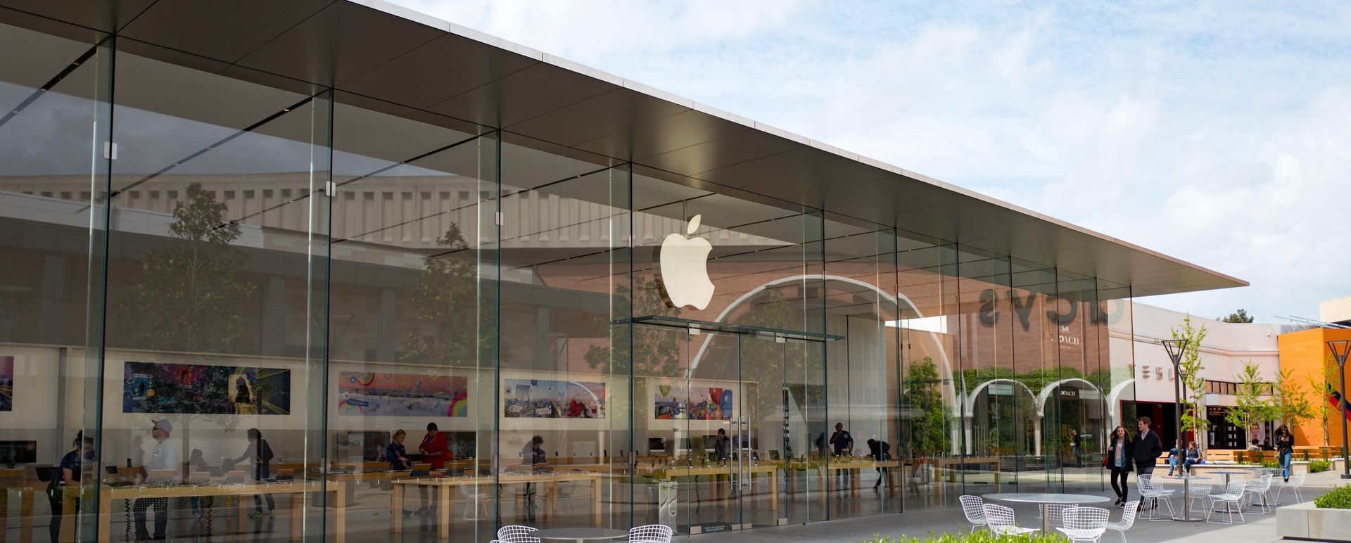 Apple Store with glass storefront and large Apple logo, people walking outside on a sunny street.