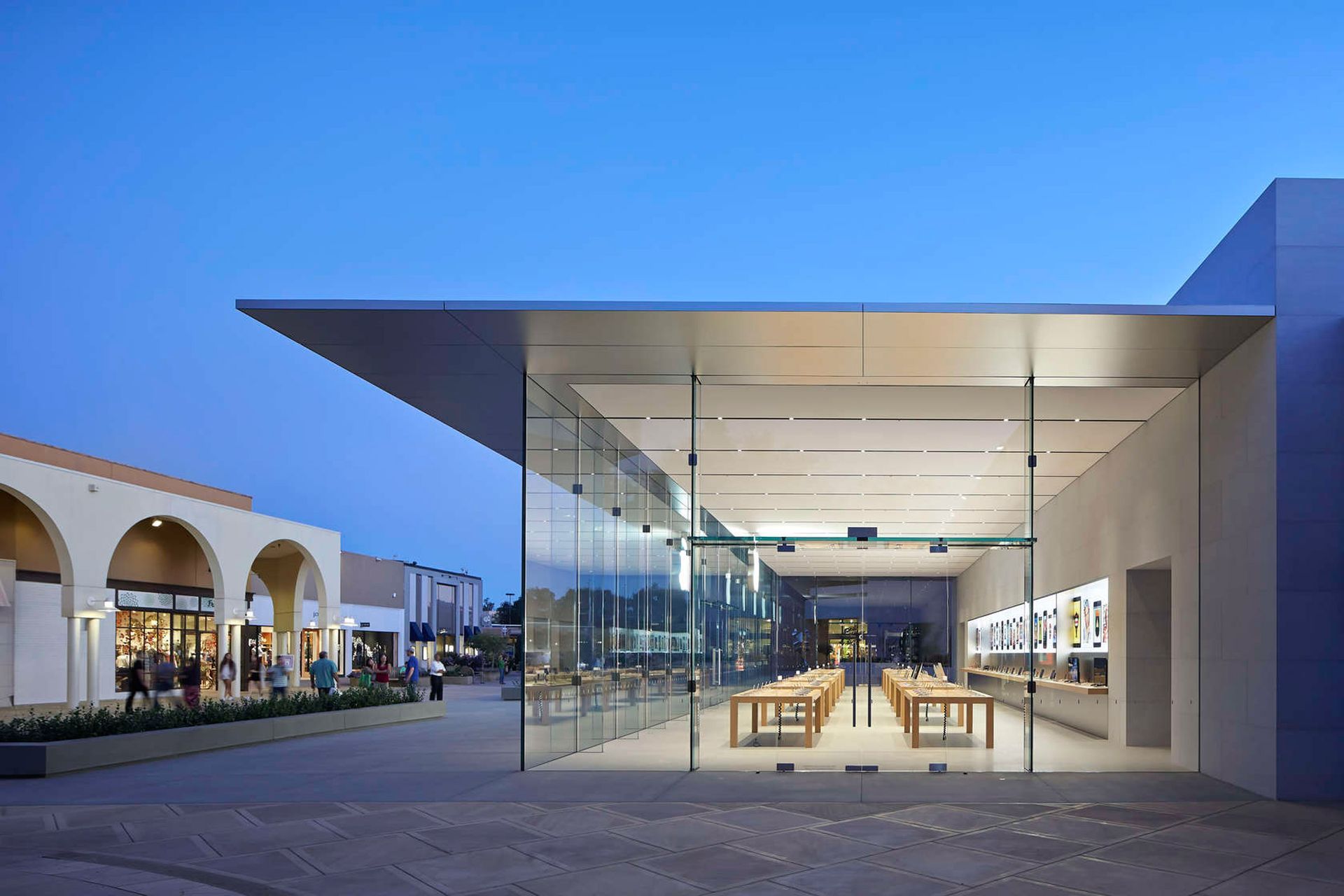 Modern glass pavilion at dusk with open seating and adjacent arched building.