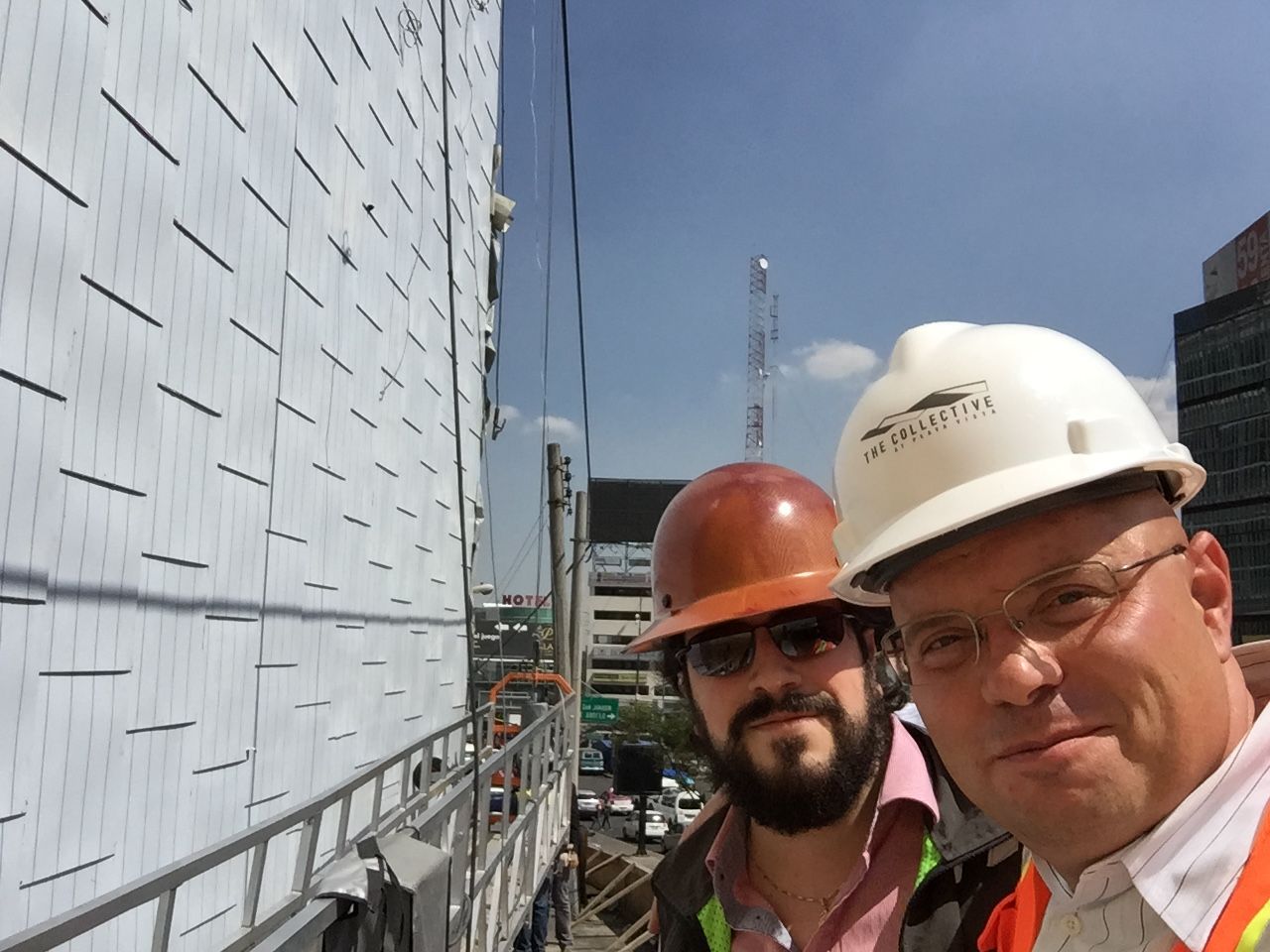 Two workers in hard hats on a rooftop beside a large white building and city skyline