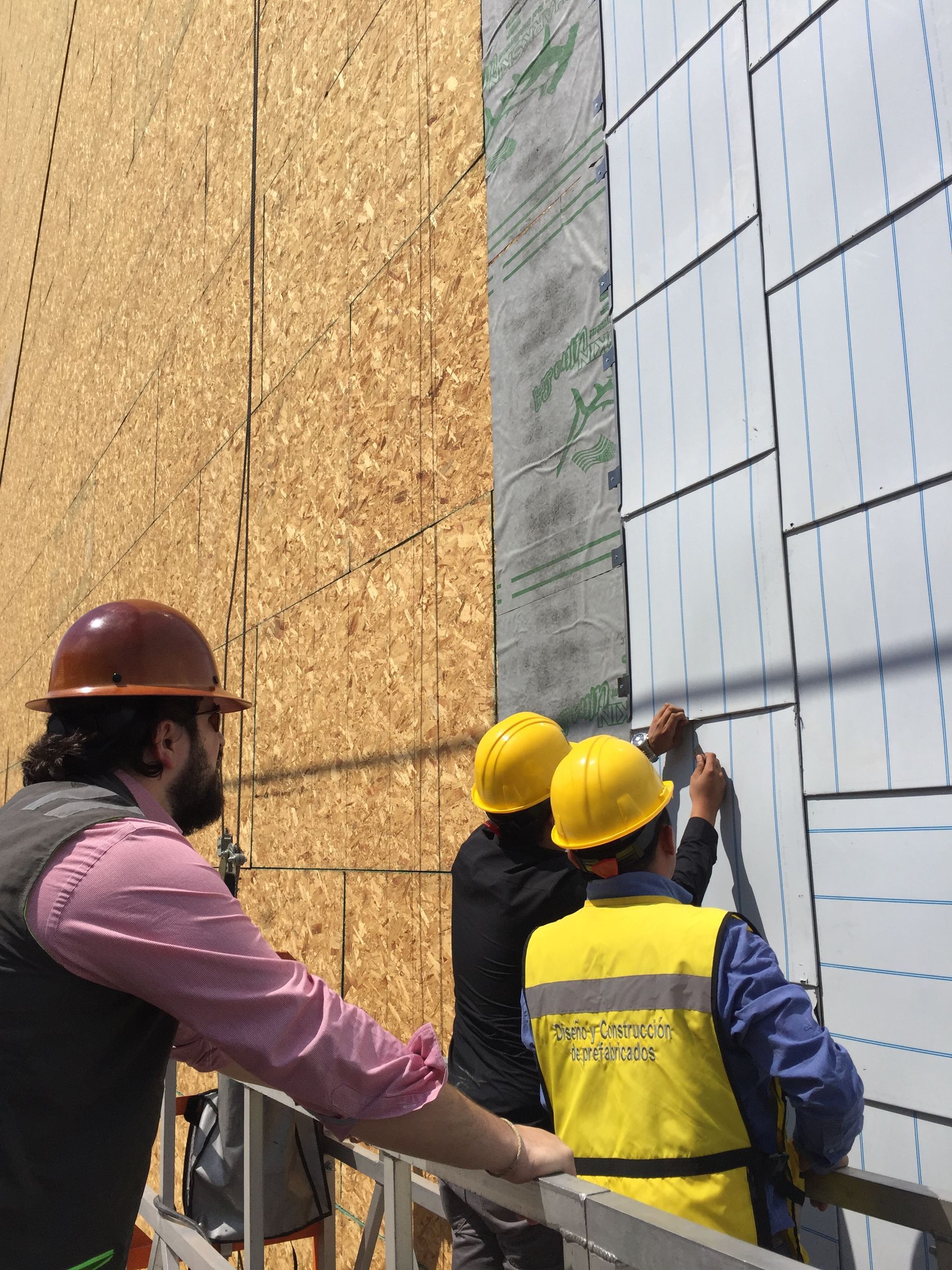 Workers in hard hats inspect a building under construction beside plywood and panel walls.