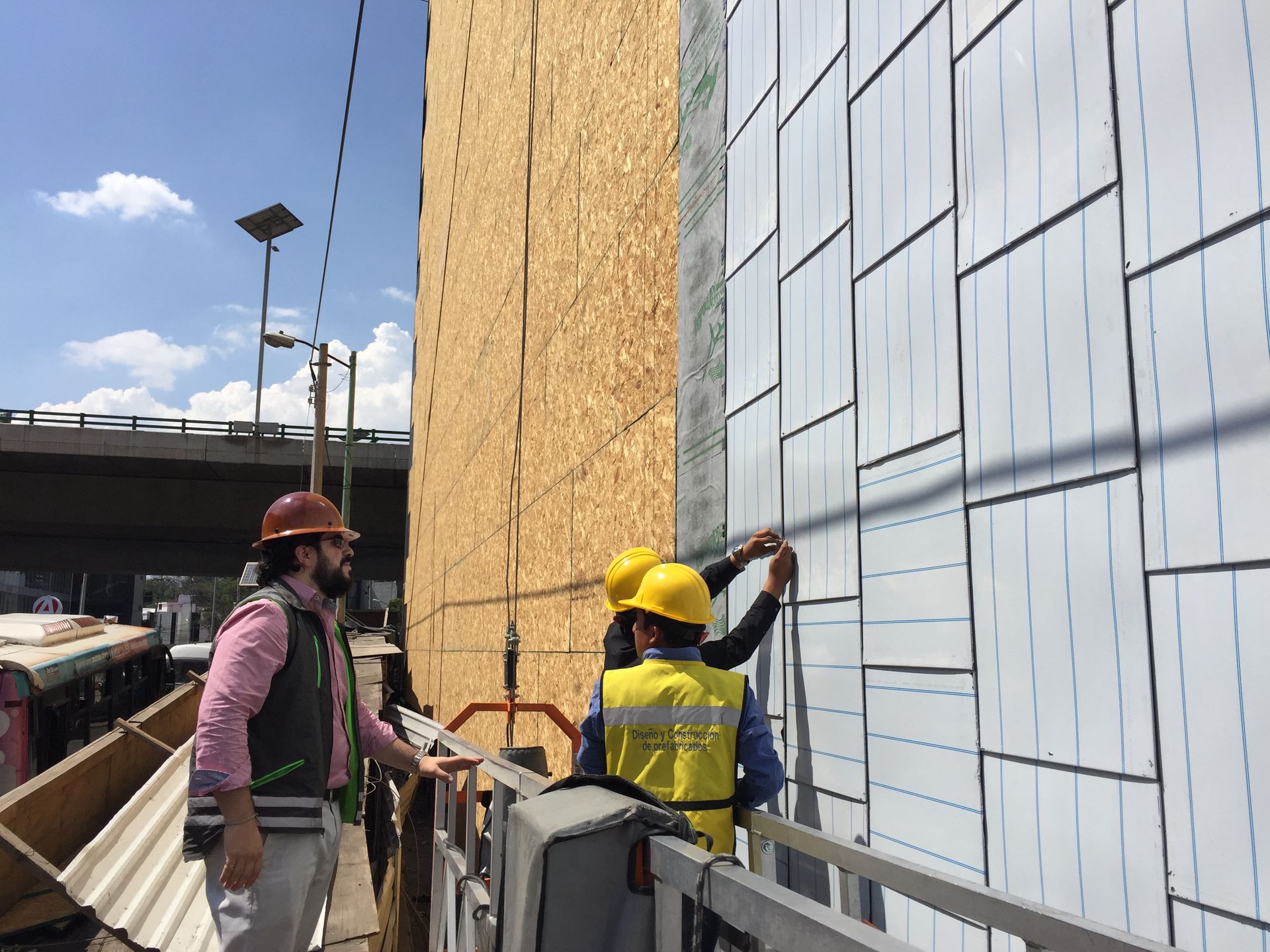 Two construction workers in hard hats inspect a wall panel installation on scaffolding.