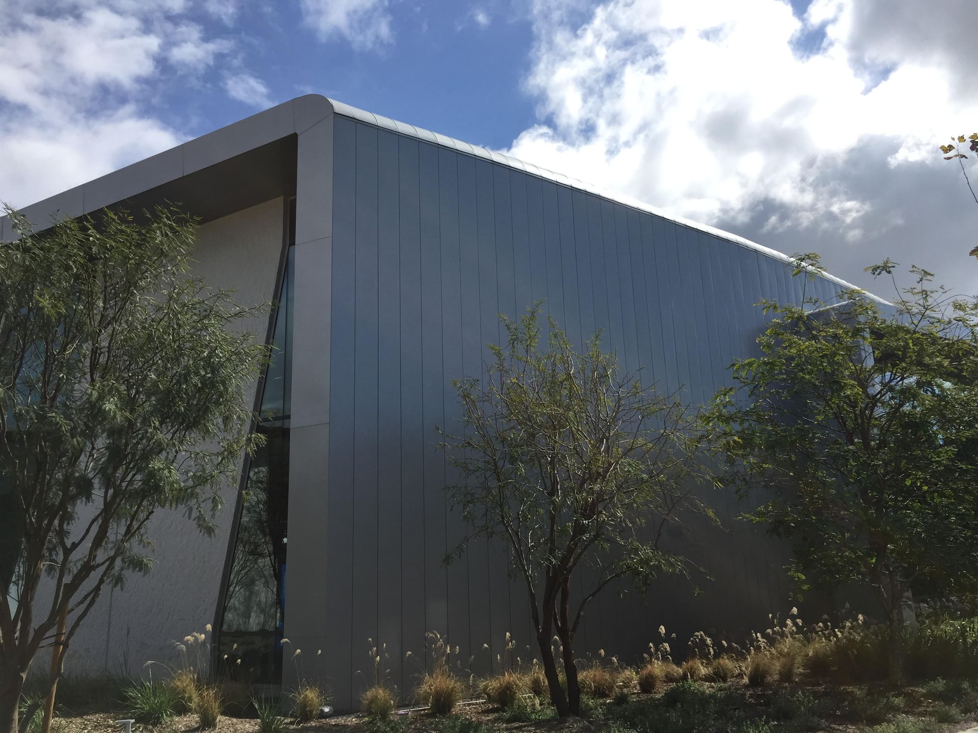 Modern gray building with a slanted facade, surrounded by trees under a partly cloudy sky