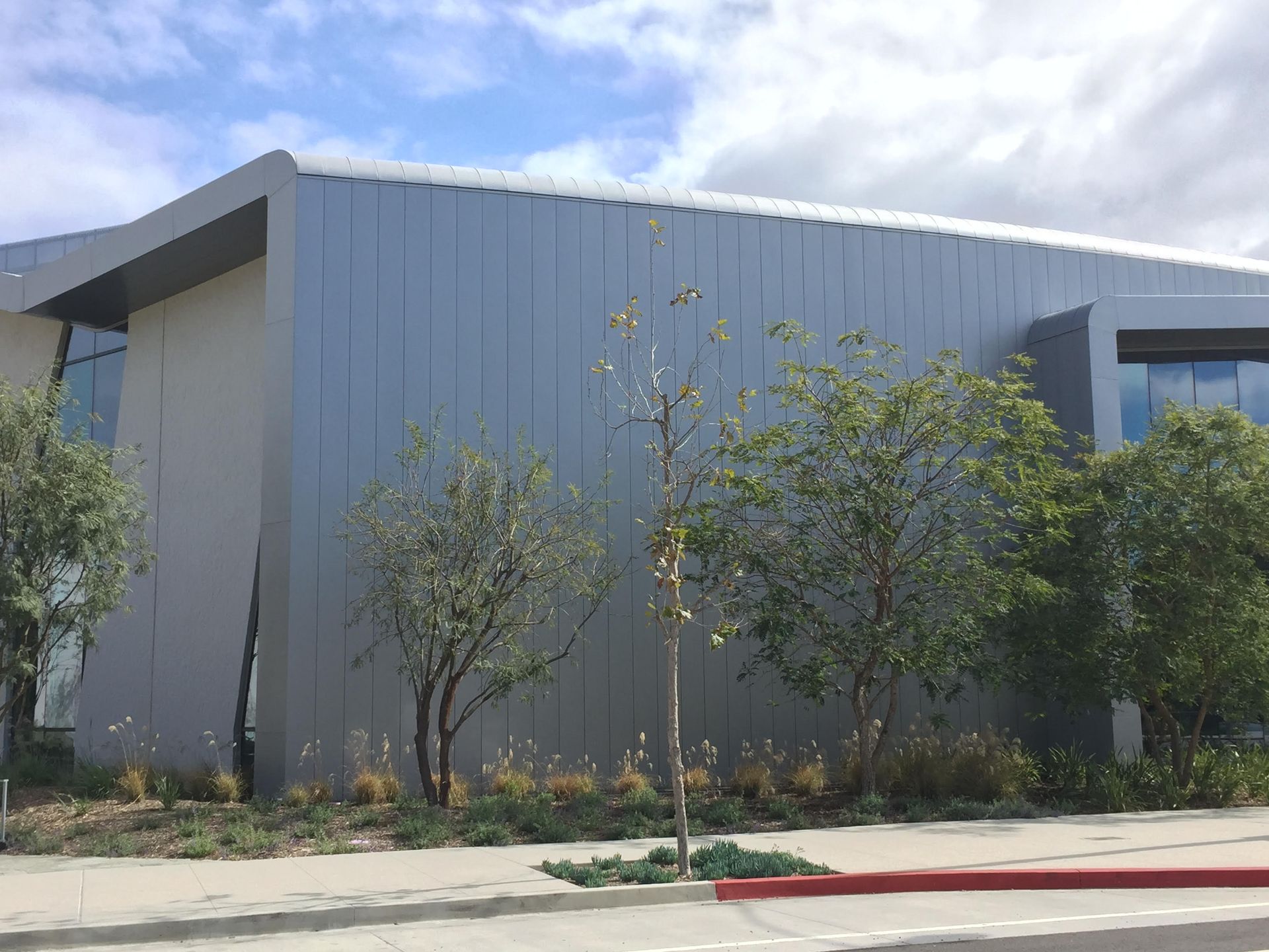 Modern gray building with trees in front and a sidewalk under a partly cloudy sky