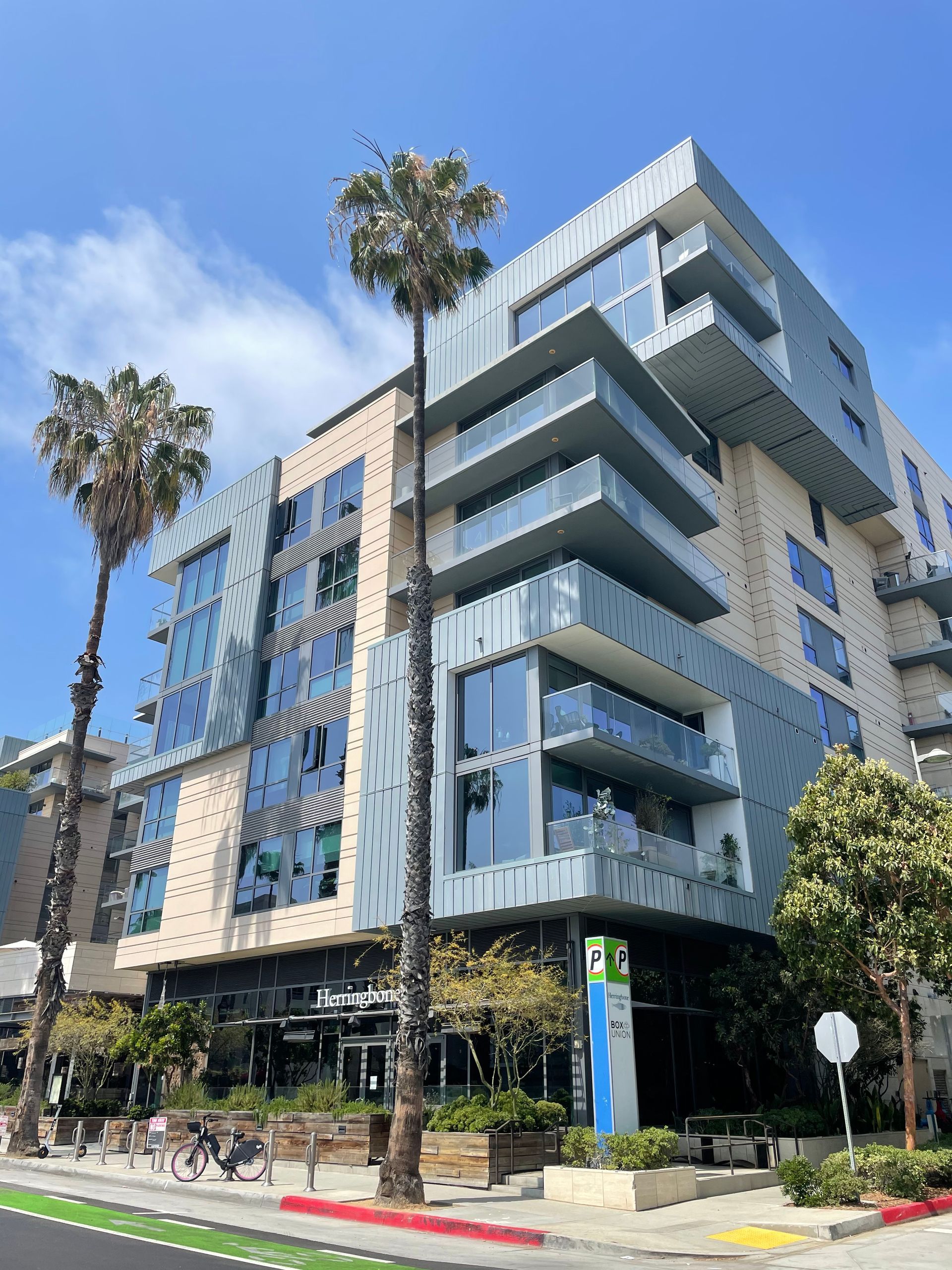 Modern blue-and-gray apartment building with balconies and palm trees along a sunny street