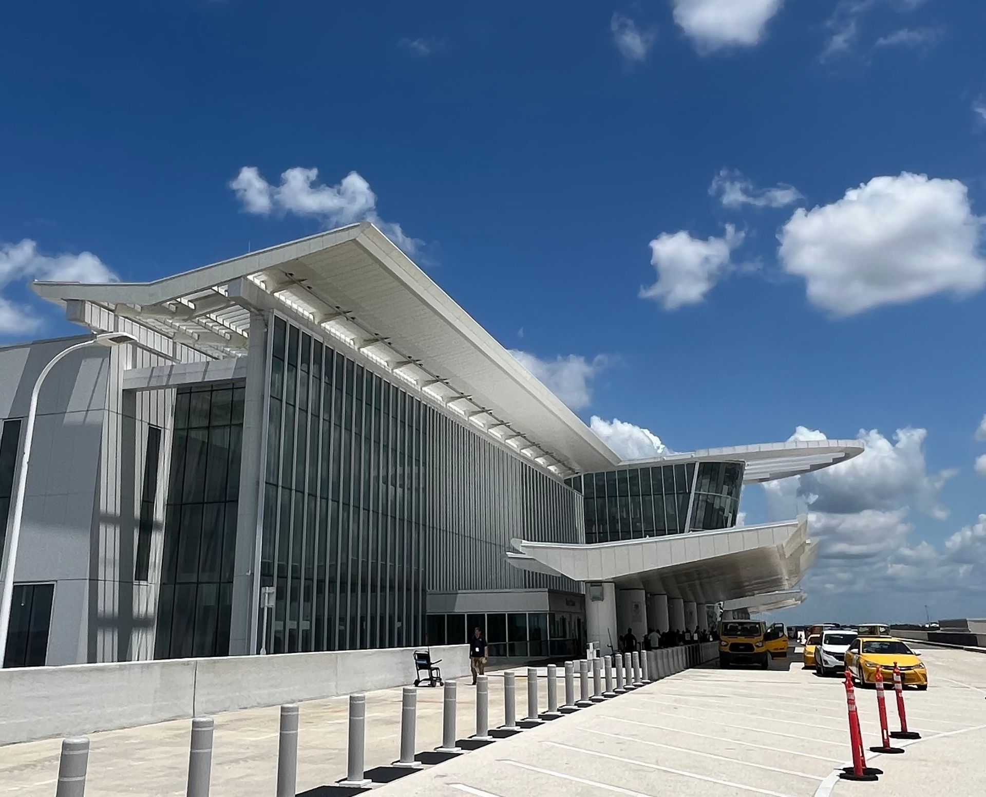 Modern glass airport terminal with a curved roof under a blue sky, taxis parked outside.