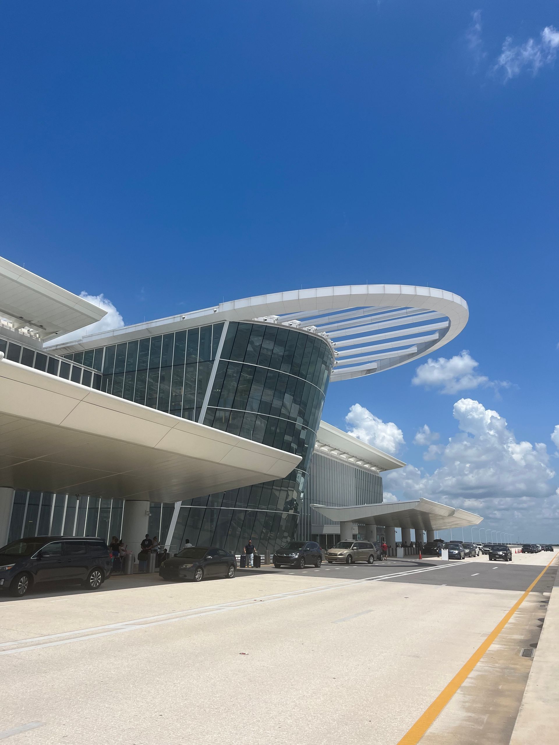 Modern airport terminal with curved glass facade under a bright blue sky, beside a wide roadway