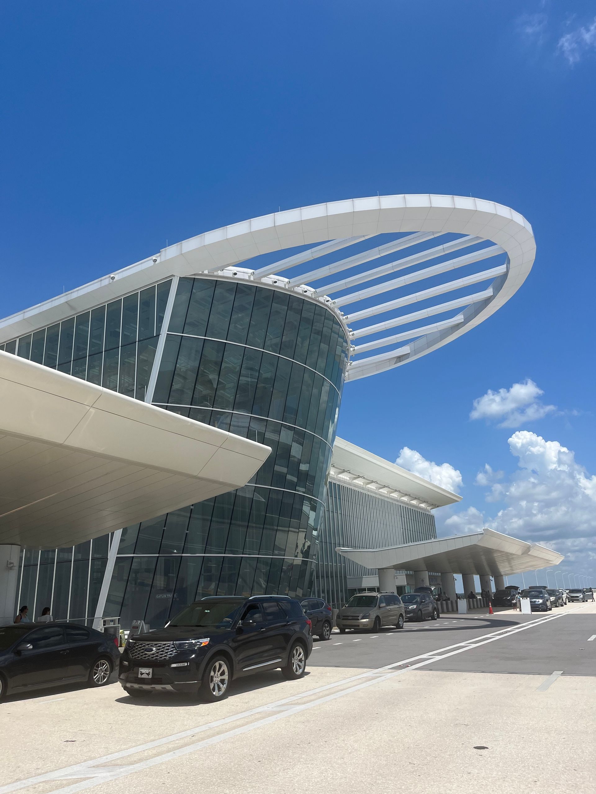 Modern airport terminal with curved glass facade, white canopy, and parked cars under a blue sky