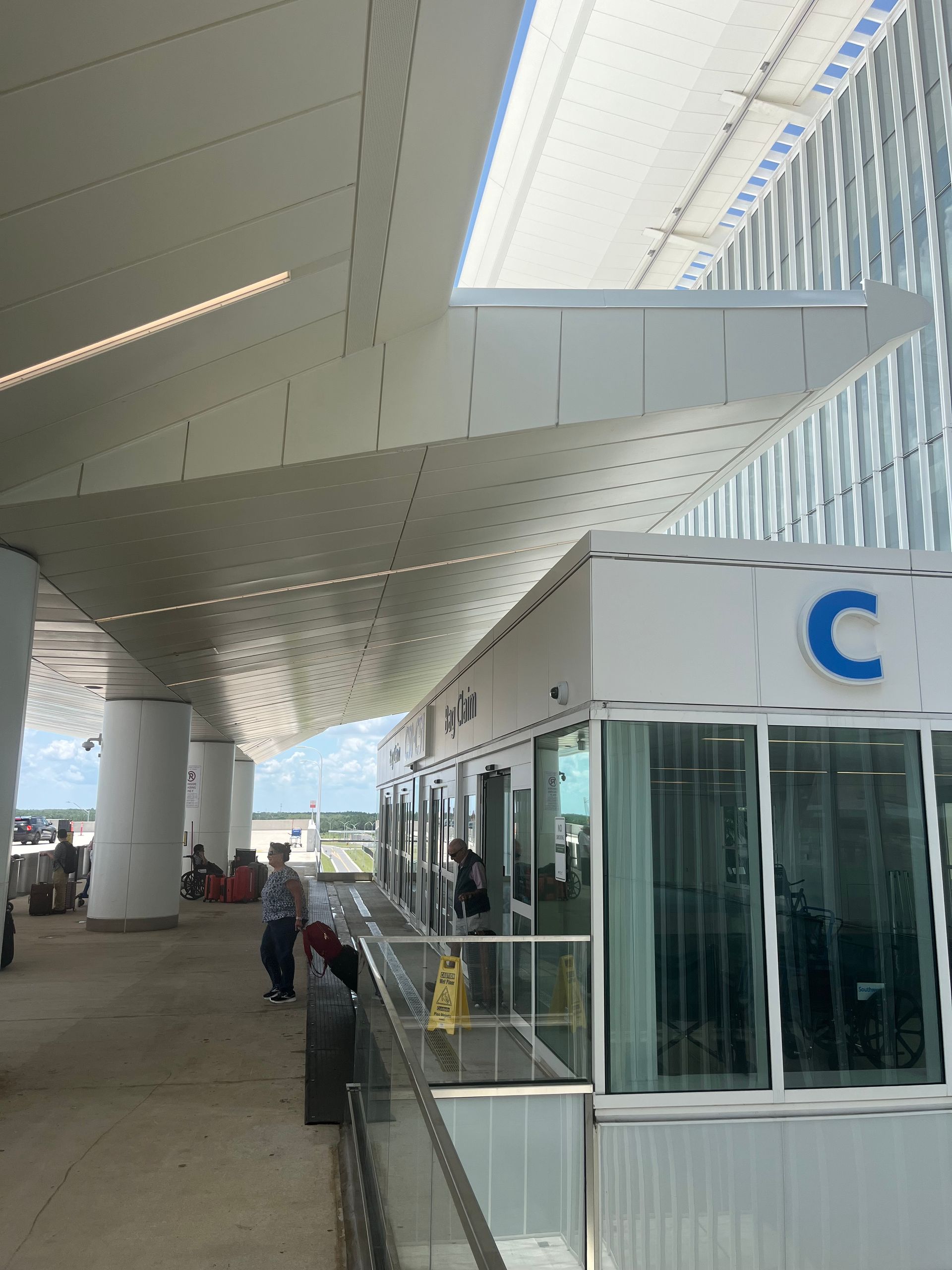 Airport curbside drop-off with white canopy, glass booth marked “C,” and a few people waiting