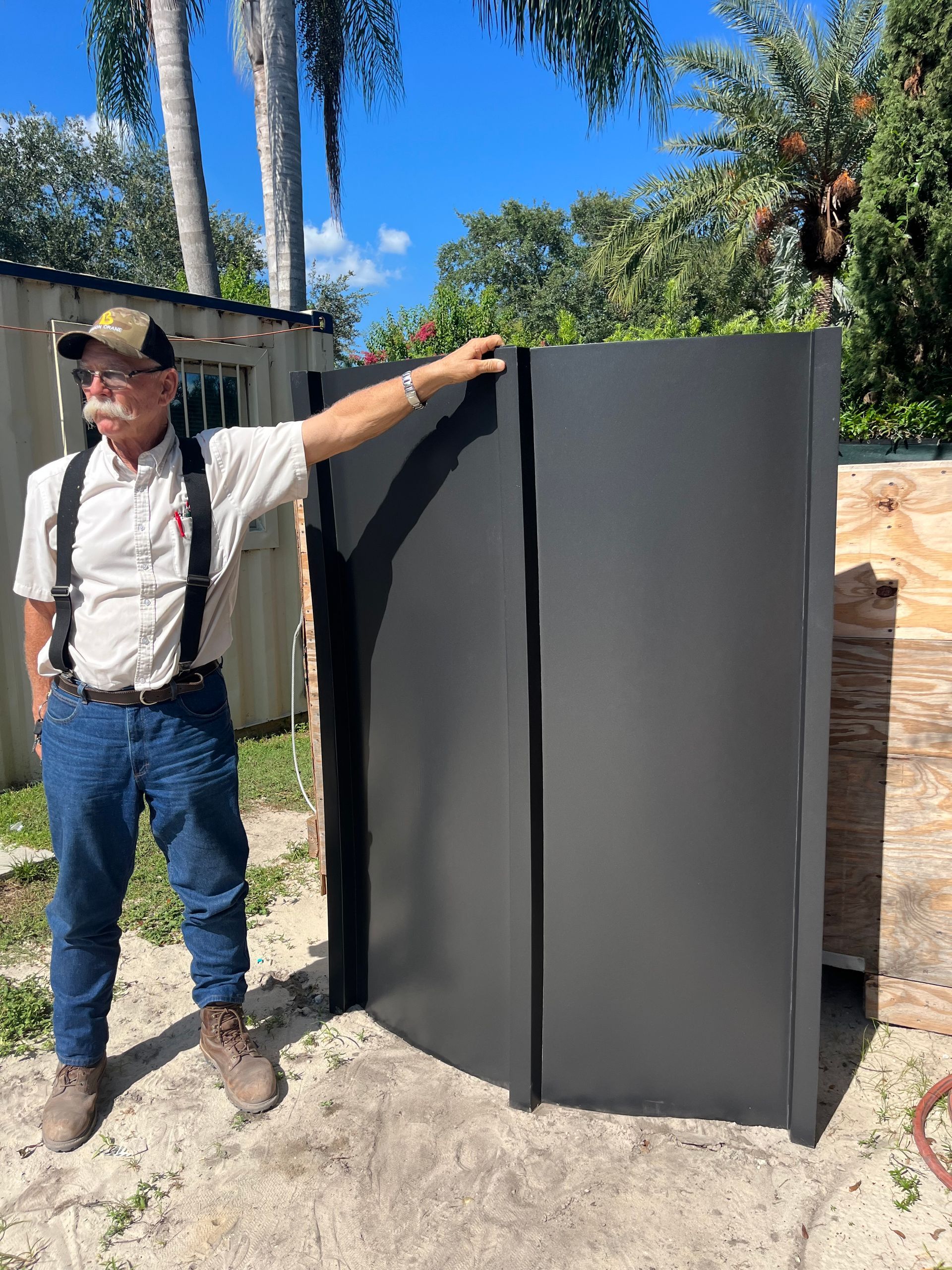 Man standing beside a large dark gray gate outdoors under a sunny blue sky