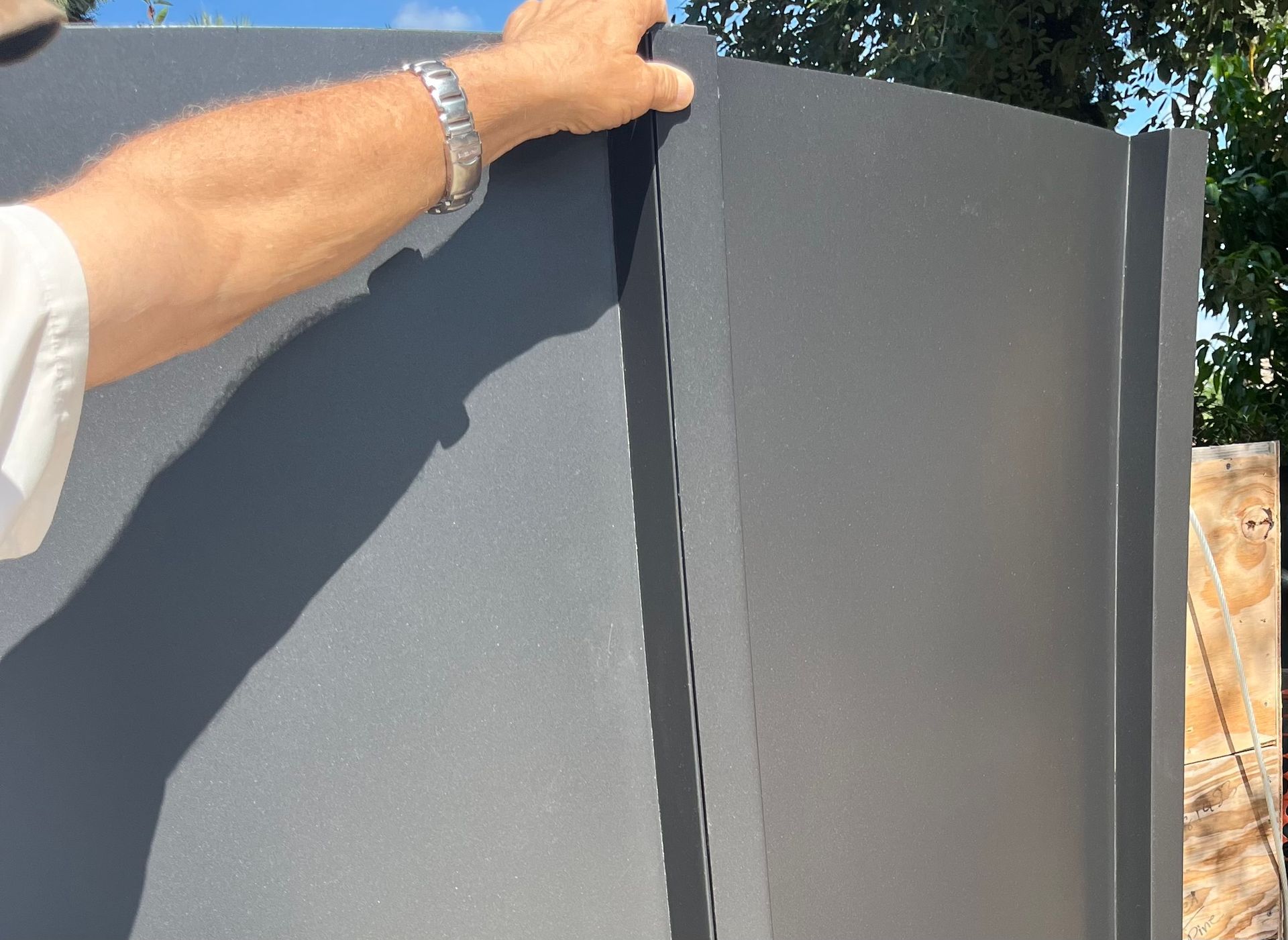 Hand holding a dark gray metal panel outdoors next to a fence