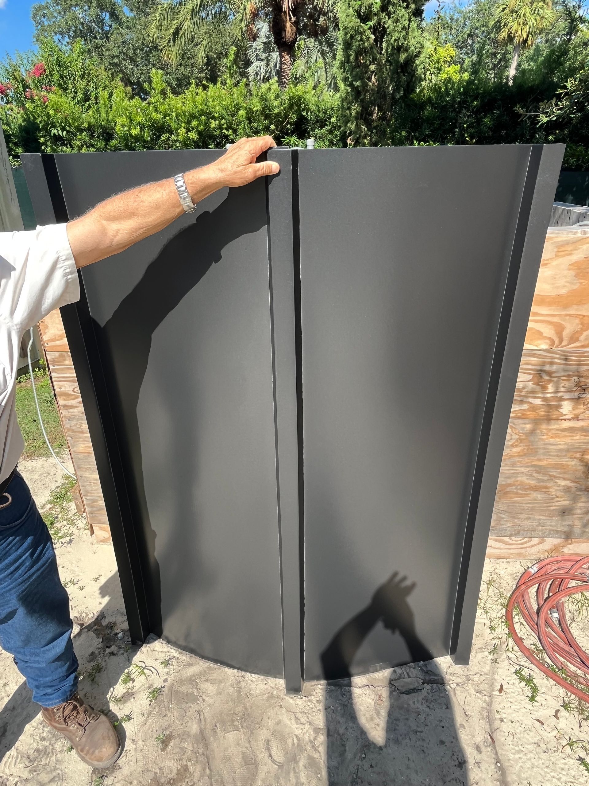 Person holding a large dark gray metal gate panel outdoors in a sunny yard.