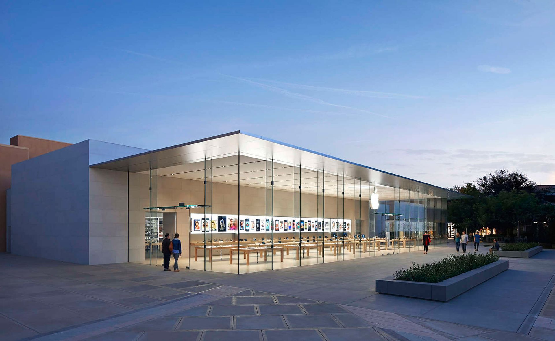 Modern glass-fronted building at dusk with lit interior and a few people outside