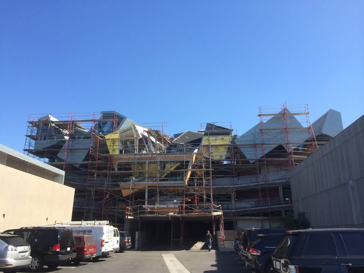 Scaffolding around a large building under construction, with parked cars in front and blue sky above