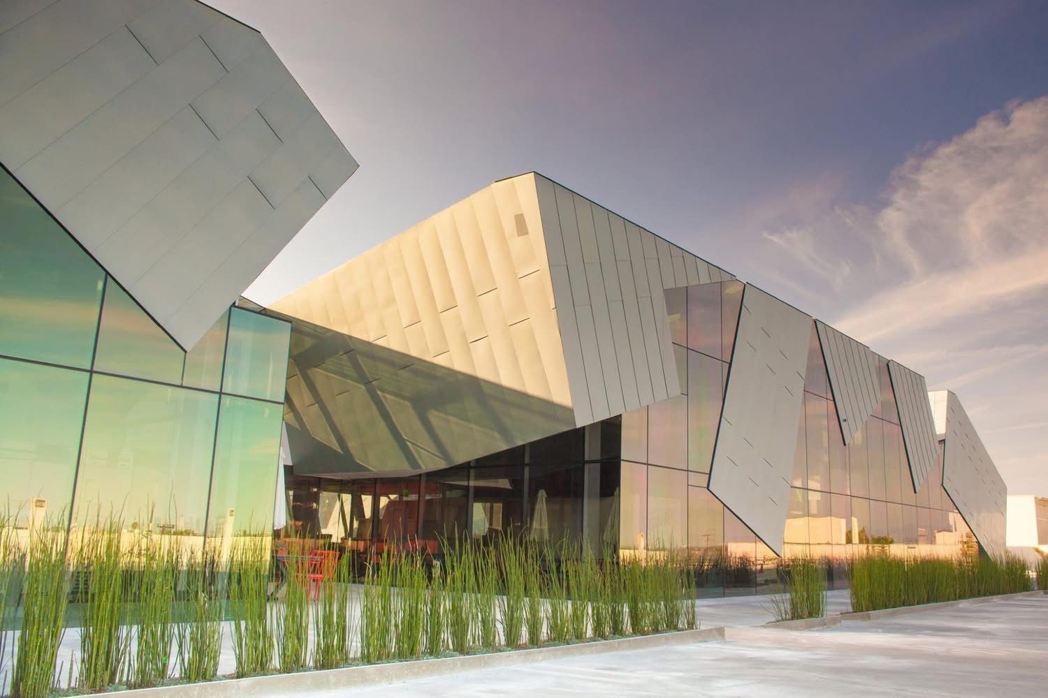 Modern angular building with glass and gold panels beside a landscaped walkway at sunset