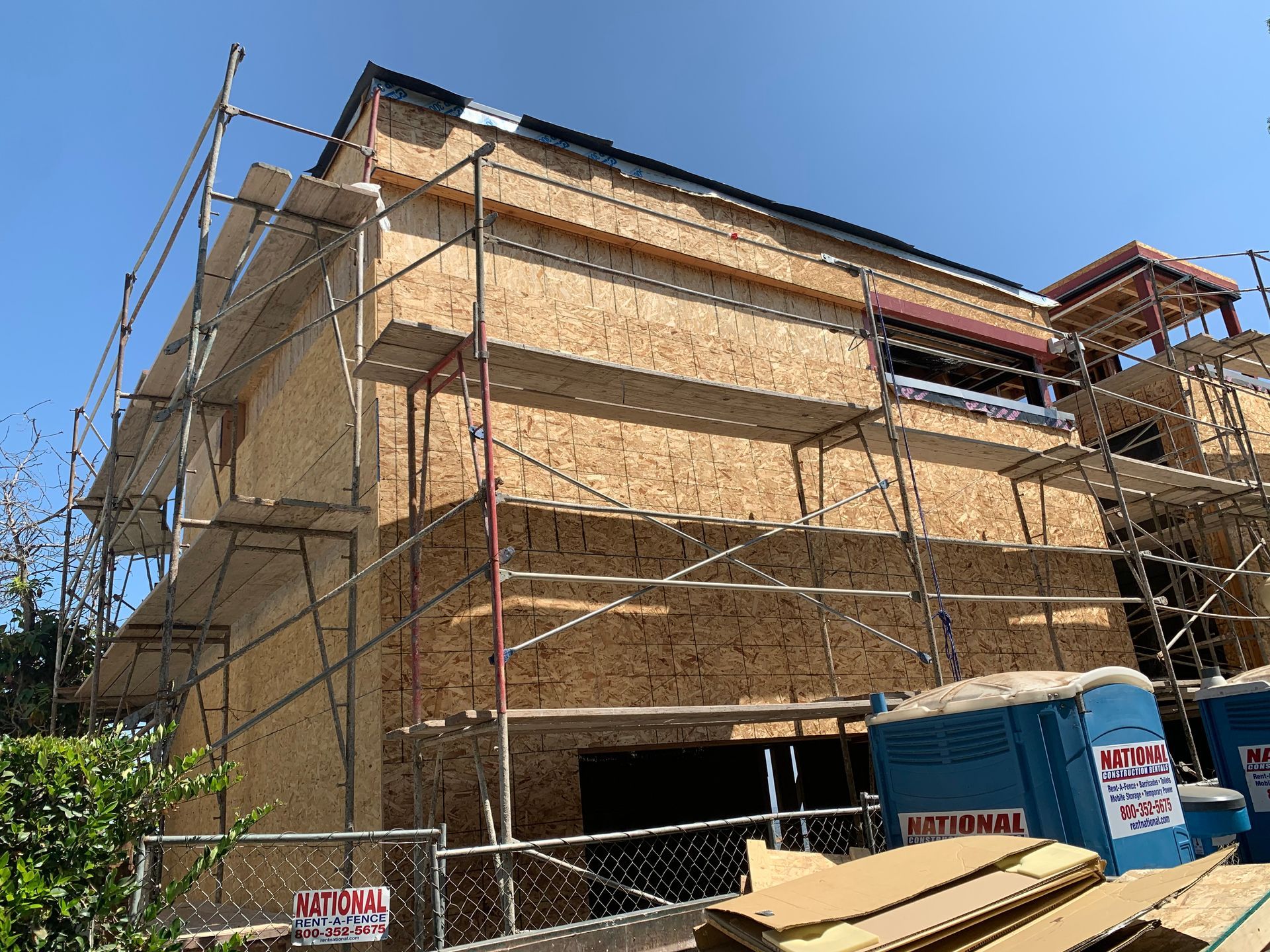 Scaffolding around a tan building under construction, with blue sky and utility trailers in front.