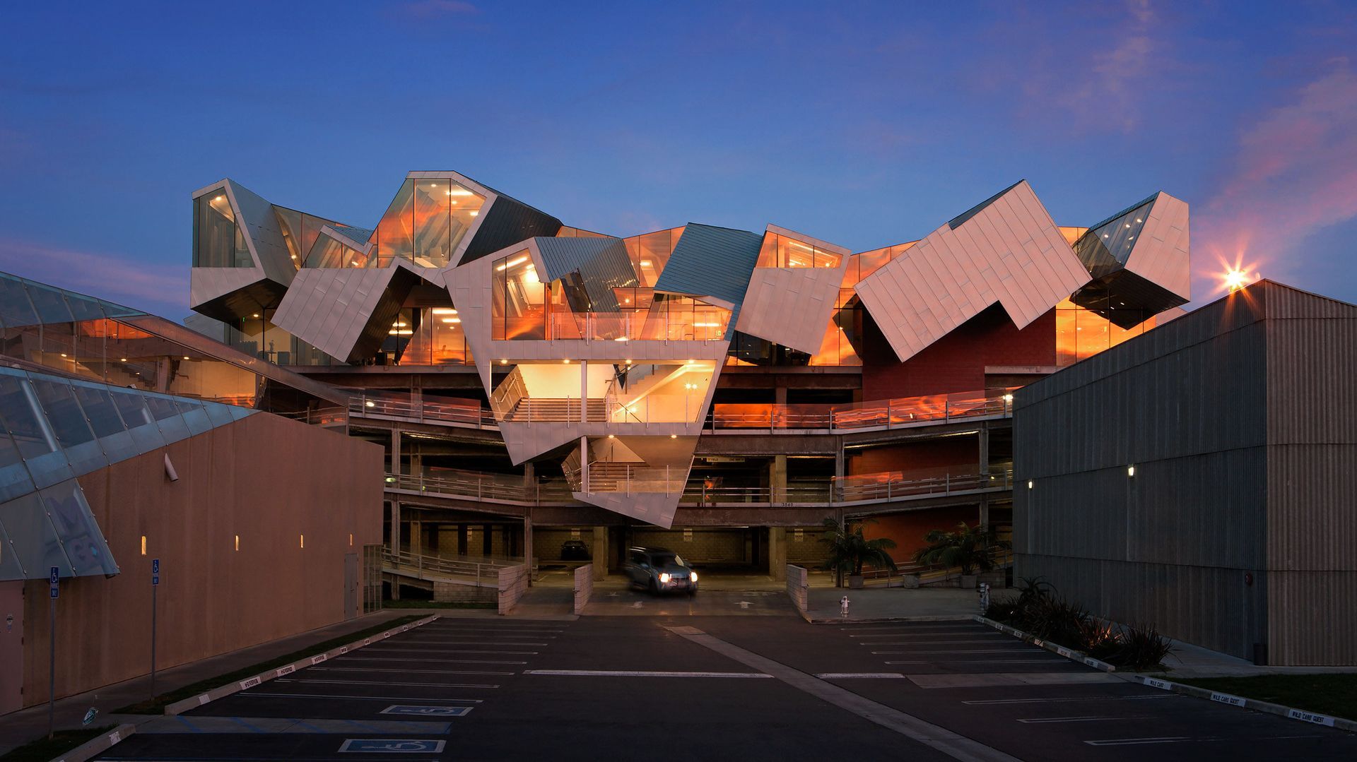Modern angular building with reflective metallic panels at dusk, lit by warm interior lights.