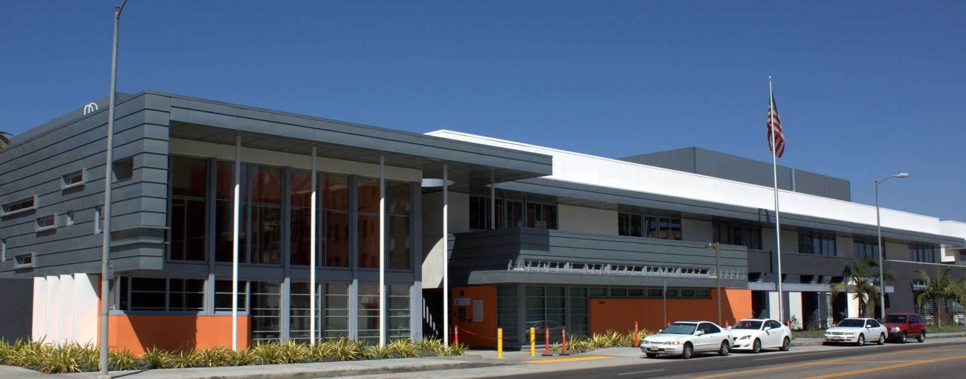 Modern building with glass entrance, American flag, and parked cars under a clear blue sky