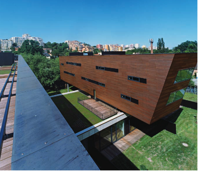 Modern angular wooden building with green roof beside a blue walkway under a clear sky