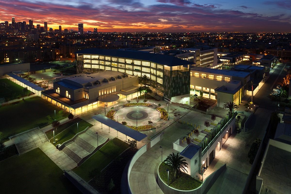 Aerial view of a modern office complex at dusk, with illuminated buildings, paths, and a city skyline beyond.