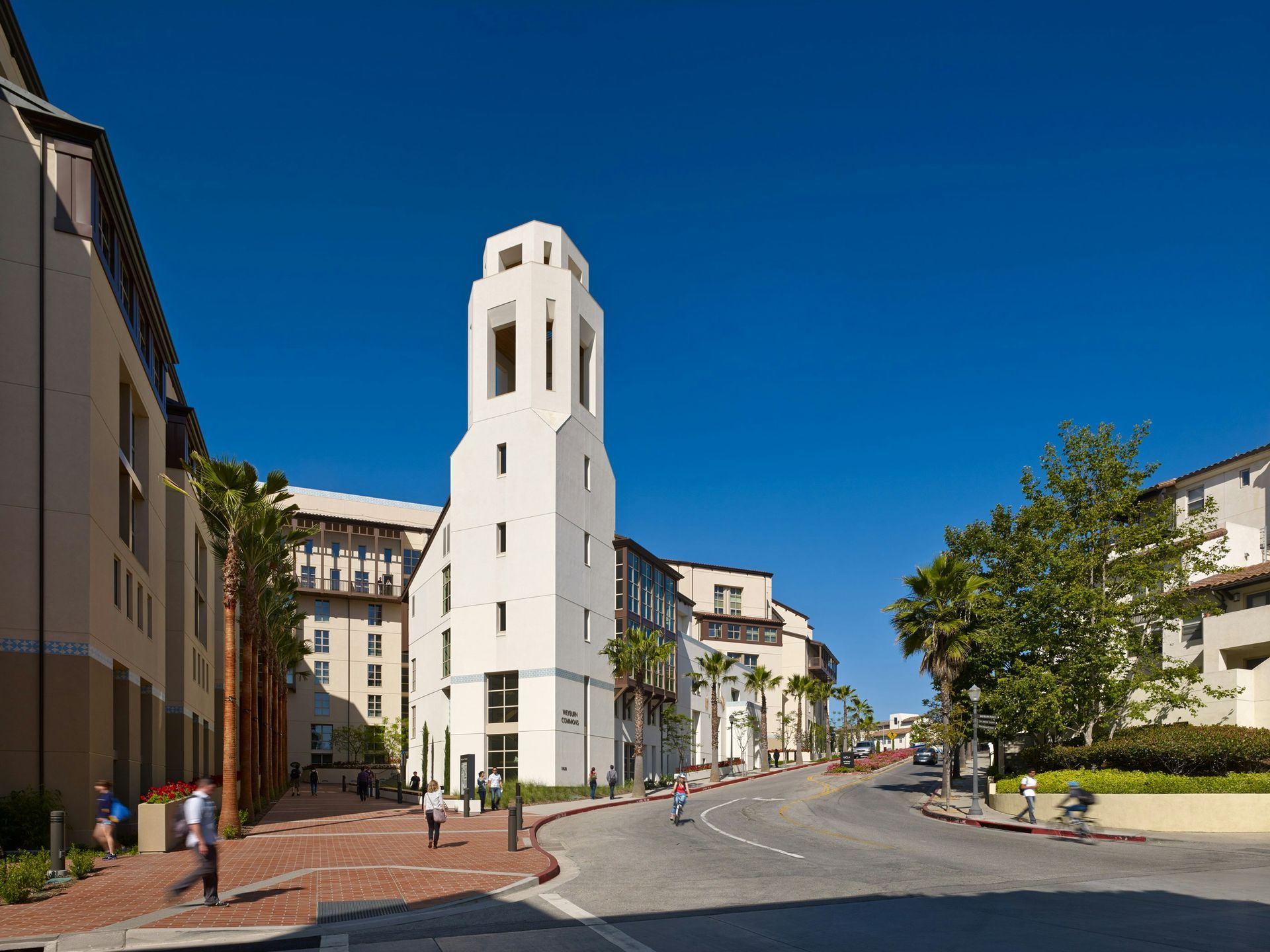 White tower-like building on a quiet street under a bright blue sky, with trees and pedestrians nearby