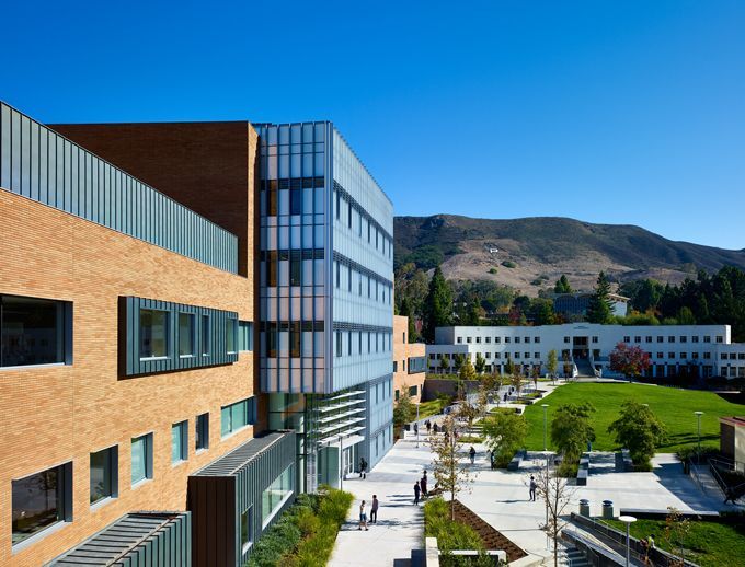 Modern campus buildings beside a green courtyard with mountains in the background under a blue sky