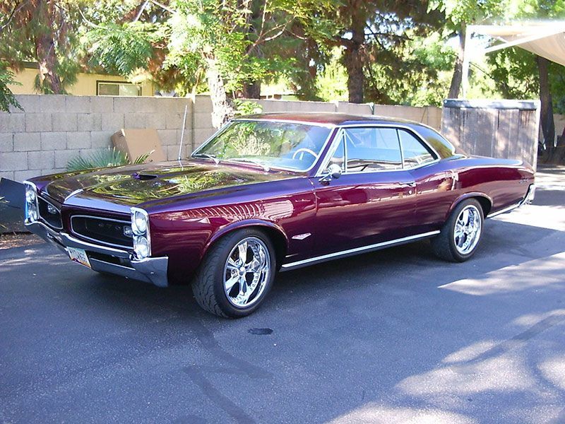Maroon classic car parked on a sunlit street beside a wall and trees