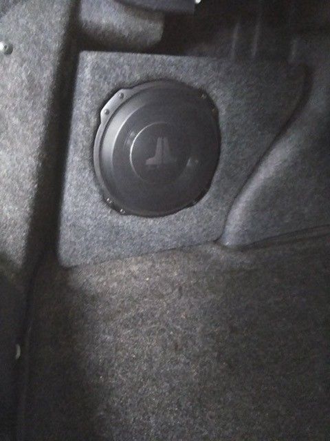 Black plastic cap with a pressure icon on a gray felt surface inside a car trunk.
