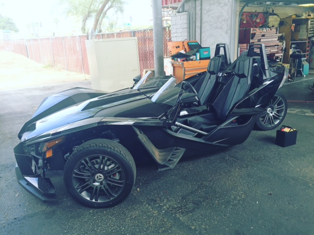 Black three-wheeled roadster with open doors parked under a covered area