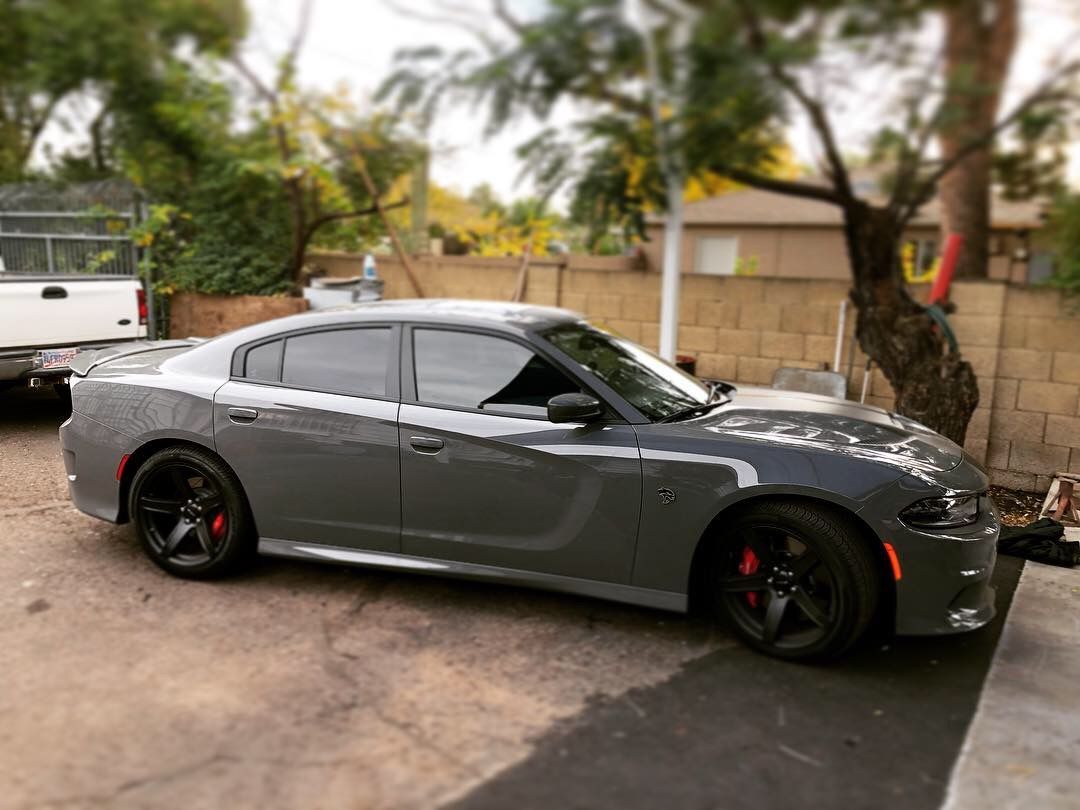 Gray sports car parked in a driveway beside a wall and trees, with a white truck in the background