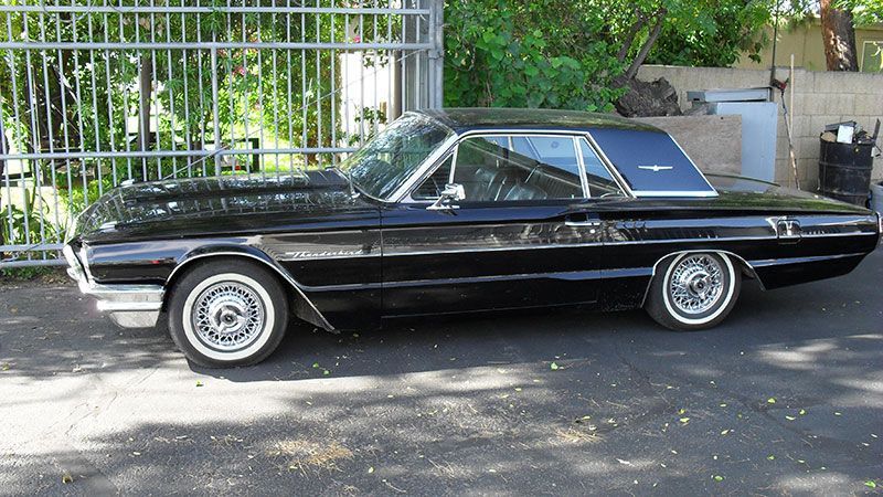 Black classic convertible parked on a street beside a metal fence and greenery.