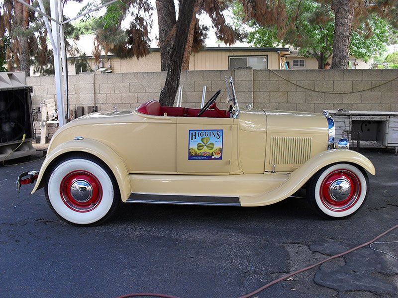 Beige vintage convertible with red wheels parked outdoors beside a wall and trees