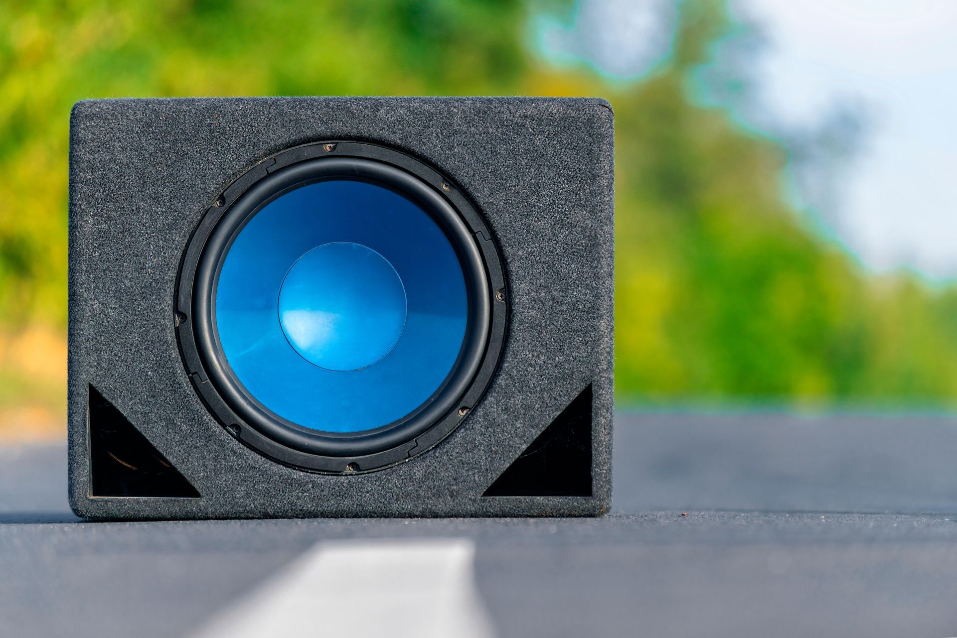 Black speaker box with a bright blue circular cone on a road, with blurred greenery in the background