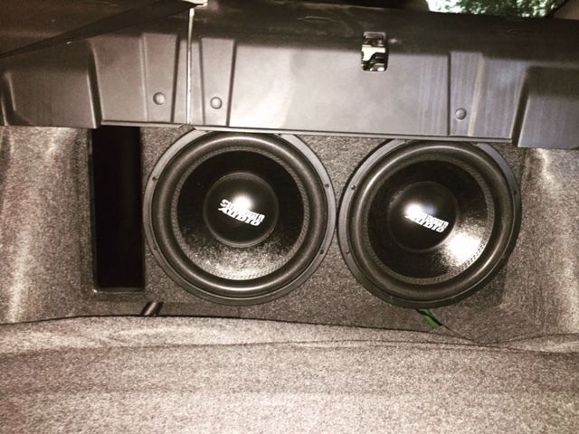 Dual black subwoofers mounted in a gray carpeted car trunk compartment
