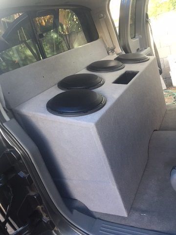 Car audio subwoofer enclosure in a vehicle cargo area, with four black speakers on a gray box.