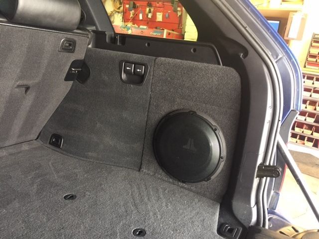 Car trunk interior with a black subwoofer in the right side panel and gray carpeted floor.