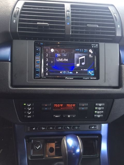 Car dashboard center console with a touchscreen stereo displaying live FM radio and climate controls below.