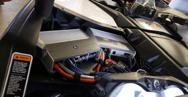 Car engine bay with exposed battery and electrical components, showing red and black cables.