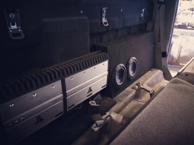 Car audio setup in a vehicle trunk, with amplifiers and two subwoofers mounted beside a folded seat.