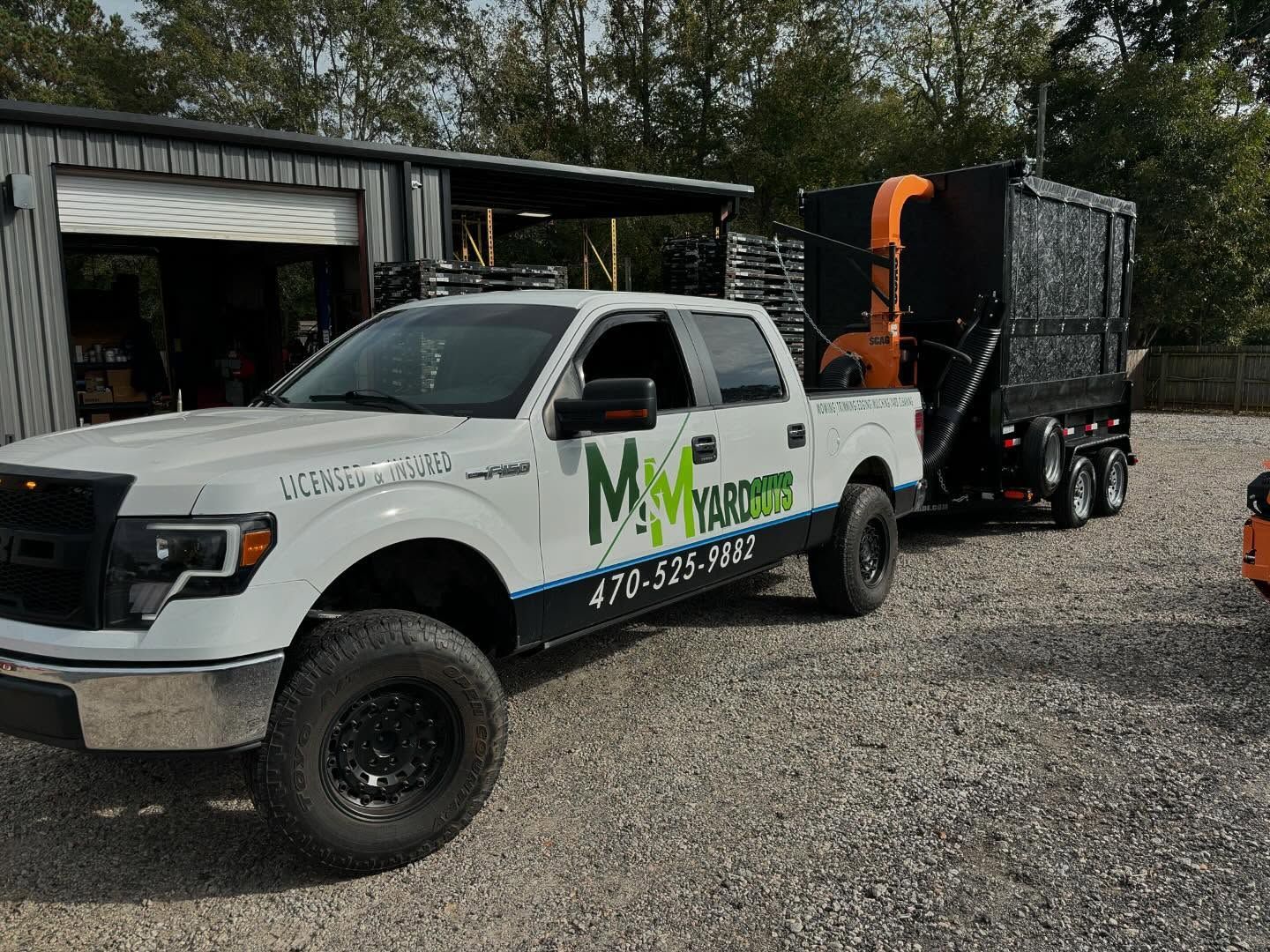 White Ford pickup truck towing a black trailer with a leaf vacuum system, parked on a gravel lot near a building.