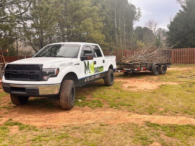 A white Ford pickup truck with MNS logo and branding, towing a utility trailer loaded with tree branches on a grass lot.