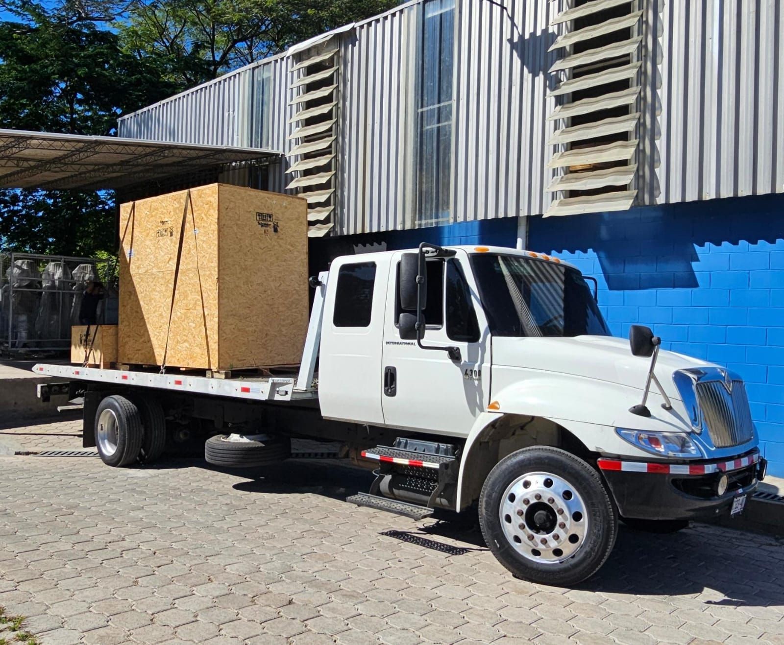 Camión de plataforma blanca que transporta cajas de madera estacionado cerca de un edificio industrial azul y plateado.
